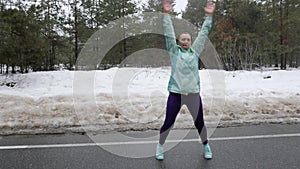 Senior old Caucasian woman warms up jumping before running in the snowy winter park. Front shot