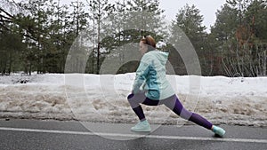 Senior old Caucasian woman warms up stretching before running in the snowy winter park. Side shot