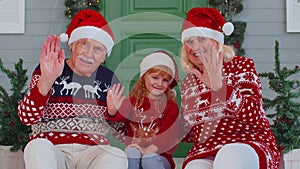 Senior old couple grandparents with grandchild girl kid waves hand hello, hi near Christmas house
