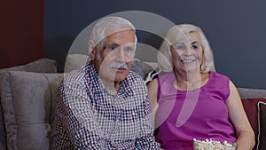 Senior old couple holding remote control talking laughing watching humor tv show sitting on sofa