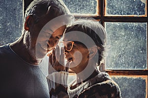 Senior old couple in love at home with snow winter outside the window. Aged woman whisper at man ear. Mature couple enjoy