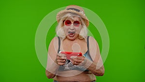 Senior old exited woman tourist in swimsuit playing video game on mobile cell phone on chroma key