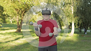 Senior old grandfather man in VR headset helmet making fitness exercises with dumbbells outdoors