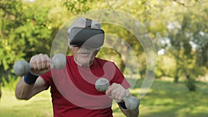 Senior old grandfather man in VR headset helmet making fitness exercises with dumbbells outdoors