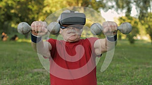 Senior old grandfather man in VR headset helmet making fitness exercises with dumbbells in park