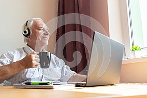 Senior old man male sit desk, home office notebook work distance freelancer listen music headphones happy call center