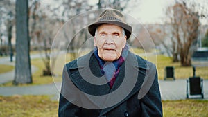 Senior old man sits in park outdoors, looks at camera zooming. Wearing coat, hat