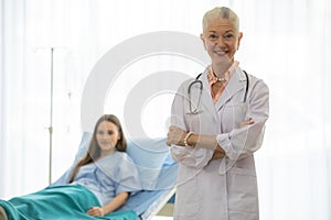 A senior older female doctor standing in an elegant pose, cross her arms, and smile with kindness with a young patient on bed blur