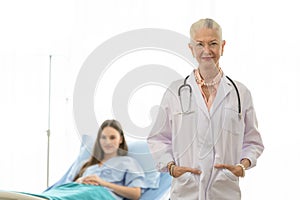 A senior older female doctor standing in an elegant pose, put hands in pocket, and smile with kindness with a young patient on bed