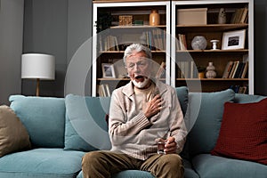 Senior older man getting chest pain in living room, heart attack holding his hand on the chest sitting on sofa at home. Health