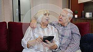 Senior pensioner couple with digital tablet pc computer at home. Resting on sofa in cozy living room