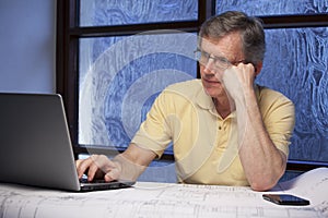 Senior pensive businessman or engineer working from home at a desk with laptop, plan and mobile phone