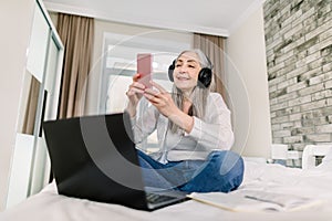 Senior peope and technologies concept. Pretty smiling retired gray haired woman in headphones, sitting on the bed, using photo