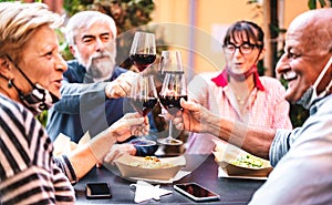 Senior people toasting wine at restaurant bar wearing opened face masks - New normal friendship concept with happy mature friends