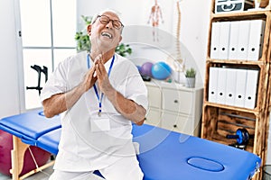 Senior physiotherapy man working at pain recovery clinic begging and praying with hands together with hope expression on face very