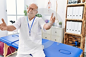 Senior physiotherapy man working at pain recovery clinic looking at the camera smiling with open arms for hug