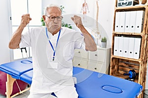 Senior physiotherapy man working at pain recovery clinic showing arms muscles smiling proud