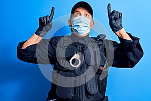Senior police man wearing surgical mask amazed and surprised looking up and pointing with fingers and raised arms