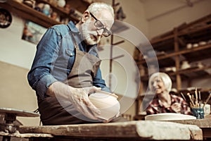 Senior potter in apron and eyeglasses examining ceramic bowl with woman working