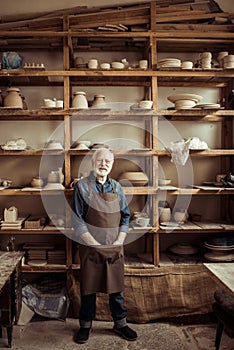 Senior potter in apron standing against shelves with pottery goods at workshop