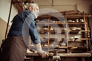 Senior potter in apron standing and leaning on table against shelves with pottery goods at workshop