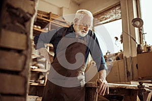 Senior potter in apron standing and leaning on table against shelves with pottery goods at workshop