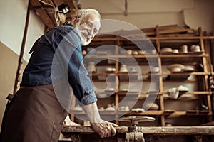 Senior potter in apron standing and leaning on table against shelves with pottery goods at workshop