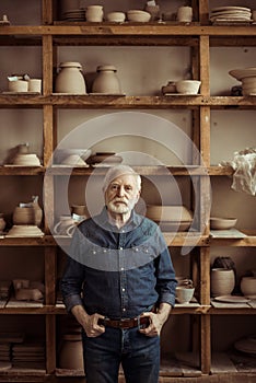 Senior potter standing against shelves with pottery goods at workshop