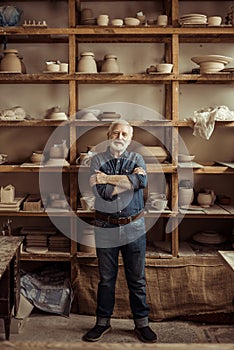 Senior potter standing against shelves with pottery goods at workshop