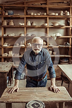 Senior potter standing and leaning on table against shelves with pottery goods at workshop