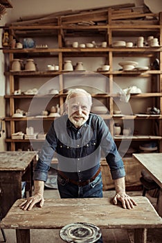 Senior potter standing and leaning on table against shelves with pottery goods at workshop