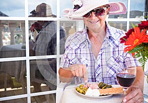 Senior pretty woman enjoying a snack with light food and a red wine glass. Healthy eating. Smiles under the pink hat. Bright light