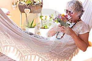 Senior pretty woman with gray hair lying on a white hammock and sniffs a bouquet of flowers. Relaxing moment outdoors on the