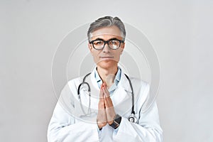 Senior professional male doctor or physician in white coat with stethoscope around neck praying, looking at camera while