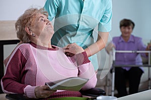 Senior reading a newspaper in common room of professional nursing home with helpful social worker supporting her