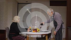 Senior retired couple having fun drinking wine and eating meal during romantic supper in kitchen