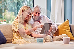 Senior Retired Couple Sitting On Sofa At Home Shopping Or Booking Holiday On Digital Tablet