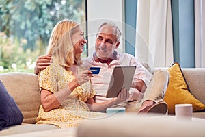 Senior Retired Couple Sitting On Sofa At Home Shopping Or Booking Holiday On Digital Tablet