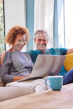 Senior Retired Couple Sitting On Sofa At Home Shopping Or Booking Holiday On Laptop