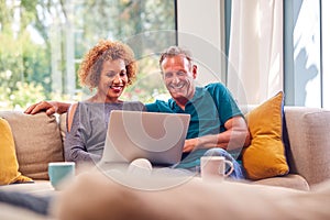 Senior Retired Couple Sitting On Sofa At Home Shopping Or Booking Holiday On Laptop