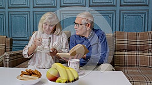 Senior retired couple talking drinking tea, reading book in modern living home room lounge together