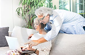 Senior retired couple using laptop computer at home on sofa - Elderly and technology concept with mature people watching shop