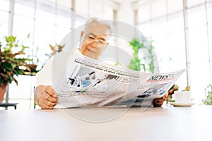 A senior retired man in casual clothes is concerned with reading a newspaper about COVID-19 news while sitting at home.