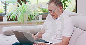 A senior retired man types on a laptop computer at home on a couch Elderly grandfather wearing eyeglasses and working on