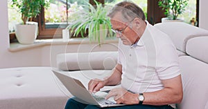 A senior retired man types on a laptop computer at home on a couch Elderly grandfather wearing eyeglasses and working on