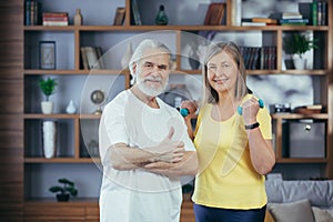 Senior retired married couple doing fitness at home, standing in living room with dumbbells and smiling looking at camera