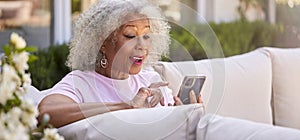 Senior Retired Woman Sitting Outside In Garden At Home Making Video Call On Mobile Phone