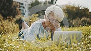 Senior retired woman using laptop in the nature while lying on the grass