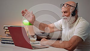 Senior smiling man pressing laptop keyboard waving talking sitting on the right indoors. Side view portrait of cheerful