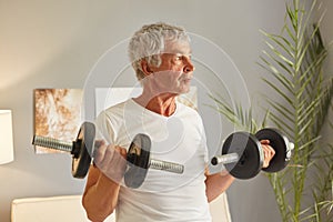 Senior sport grey-haired mature man using barbels foe training biceps and triceps having home workout wearing casual white T-shirt